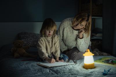 A woman and child sitting on a bed. The child is drawing on a clipboard. The room is completely dark except for light coming from battery-powered lantern on the bed next to them.