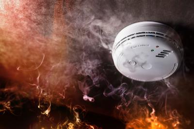 A close-up of a smoke alarm during a fire, with smoke surrounding the alarm and flames glowing underneath.