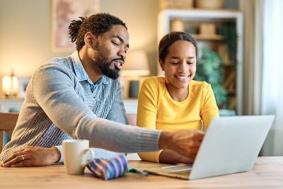 Father and child sitting at a table, talking about what is on the laptop in front of them.